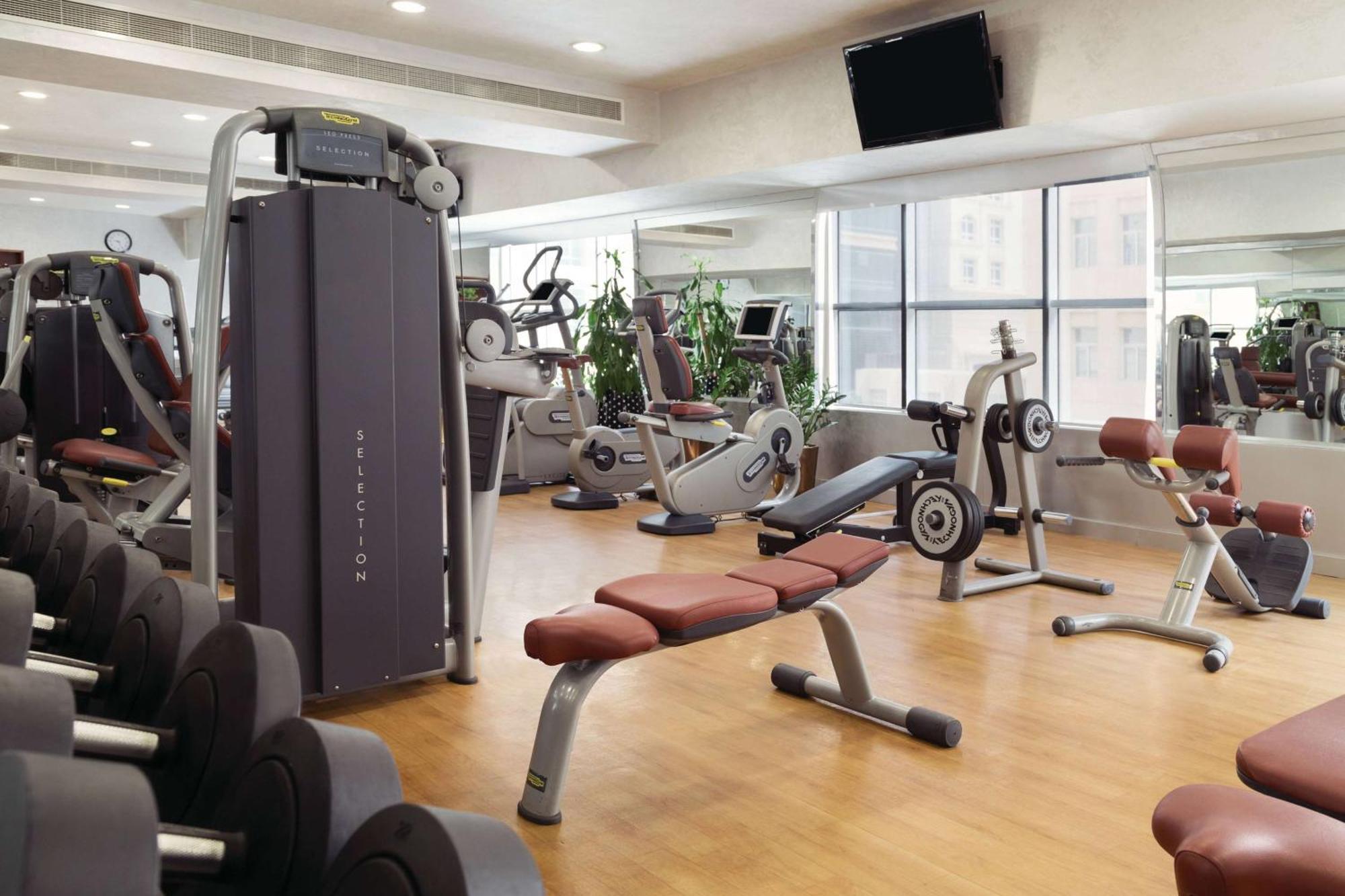Grand Regency Doha, Trademark Collection By Wyndham Екстер'єр фото The photo shows a fitness gym with various exercise equipment. There are weight machines, benches, and cardio machines such as stationary bikes and treadmills. The space has large windows allowing natural light to enter, and there are mirrors on the 