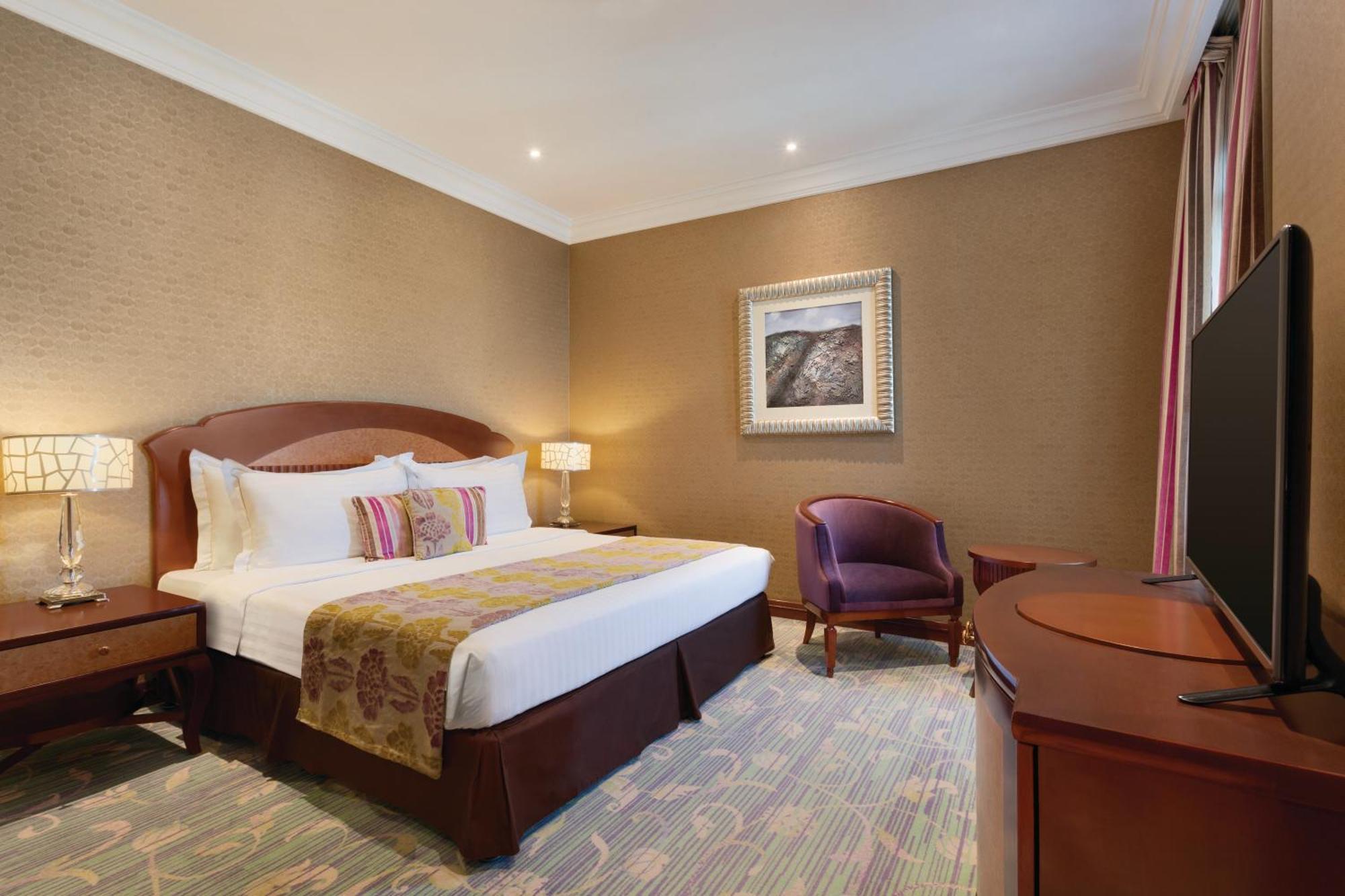 Grand Regency Doha, Trademark Collection By Wyndham Екстер'єр фото The image shows a hotel room that features a cozy and elegant design. There's a large bed with white sheets and colorful decorative pillows. Next to the bed, there are nightstands with lamps. A comfortable armchair is positioned in the corner, and a 