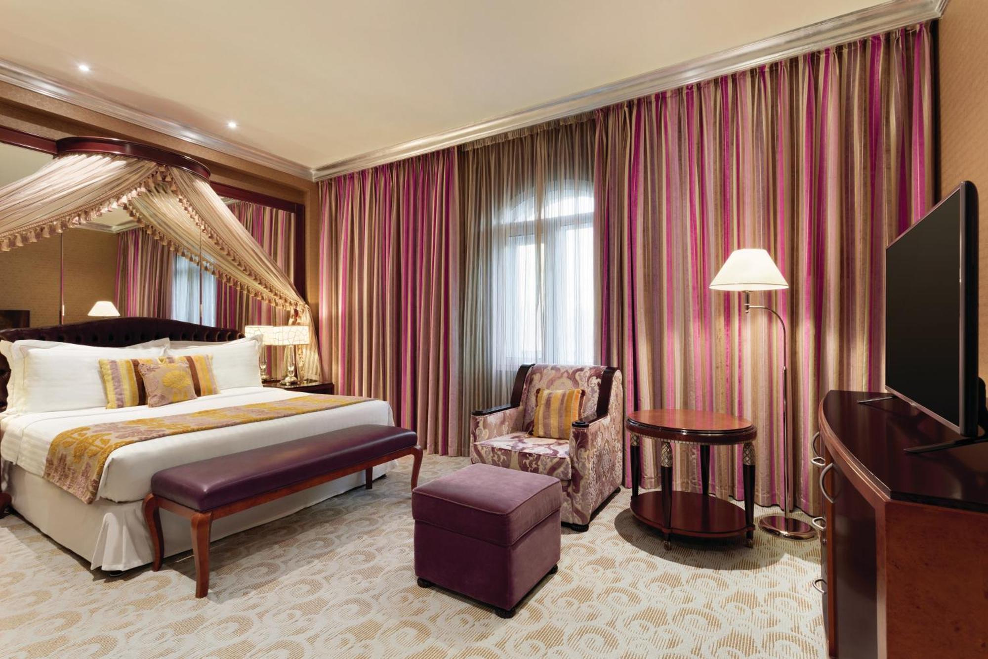 Grand Regency Doha, Trademark Collection By Wyndham Екстер'єр фото The photo shows a stylish hotel room. It features a large bed with decorative pillows and a draped canopy above it. The room has elegant striped curtains in shades of pink and purple. There is a small seating area with a plush chair and ottoman, alon