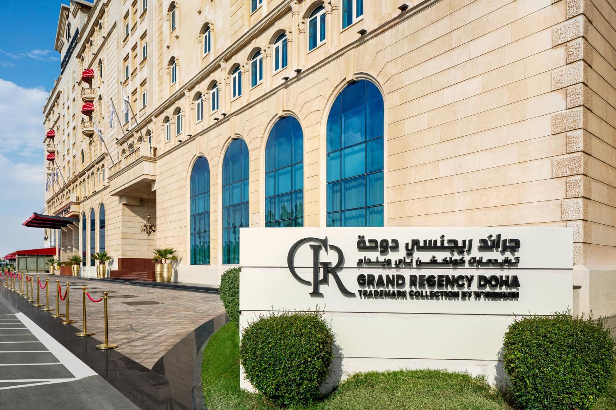 Grand Regency Doha, Trademark Collection By Wyndham Екстер'єр фото The photo shows the entrance of the Grand Regency Doha, a luxury hotel. The building features a distinctive architectural style with large arched windows. There is a prominent sign featuring the hotel's name in both English and Arabic. The surroundin
