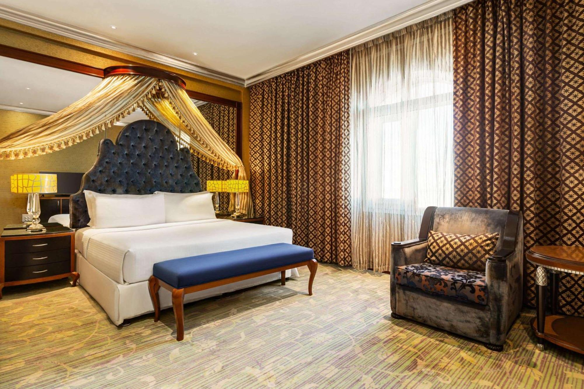 Grand Regency Doha, Trademark Collection By Wyndham Екстер'єр фото The photo shows a luxurious hotel room featuring a large bed with an intricately designed headboard and elegant bedding. Above the bed, there are decorative drapes that add a touch of sophistication. In front of the bed, there is a blue bench. To the
