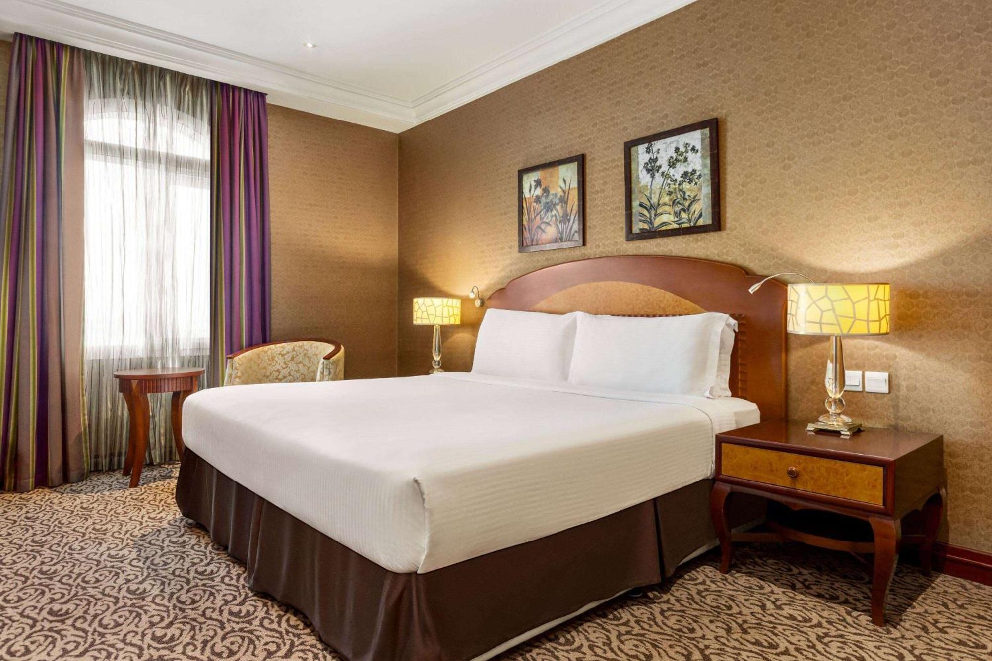 Grand Regency Doha, Trademark Collection By Wyndham Екстер'єр фото The photo displays a comfortable hotel room featuring a large bed with white linens and a brown bed skirt. The room has a warm color palette, with textured brown walls and patterned carpet. There are two bedside lamps providing soft lighting next to 