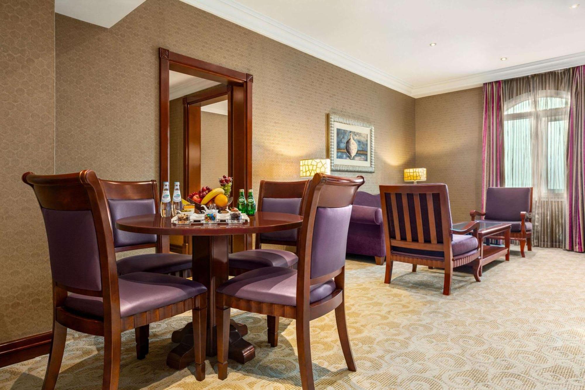 Grand Regency Doha, Trademark Collection By Wyndham Екстер'єр фото The photo depicts a well-furnished hotel room or lounge area. In the foreground, there is a round table surrounded by four purple upholstered chairs, with a selection of fruits and beverages on the table. To the right, there are two wooden armchairs,